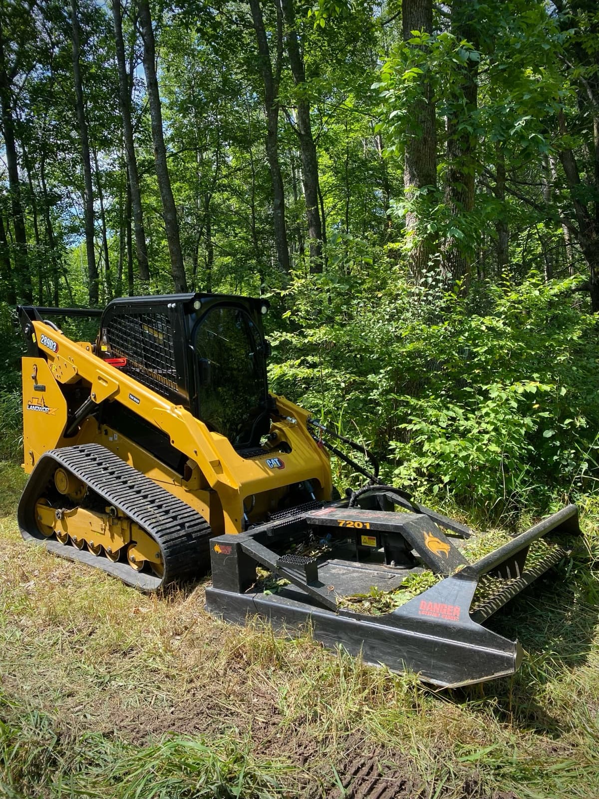 Compact track loader with rotary brush cutter mowing heavy brush at the tree line