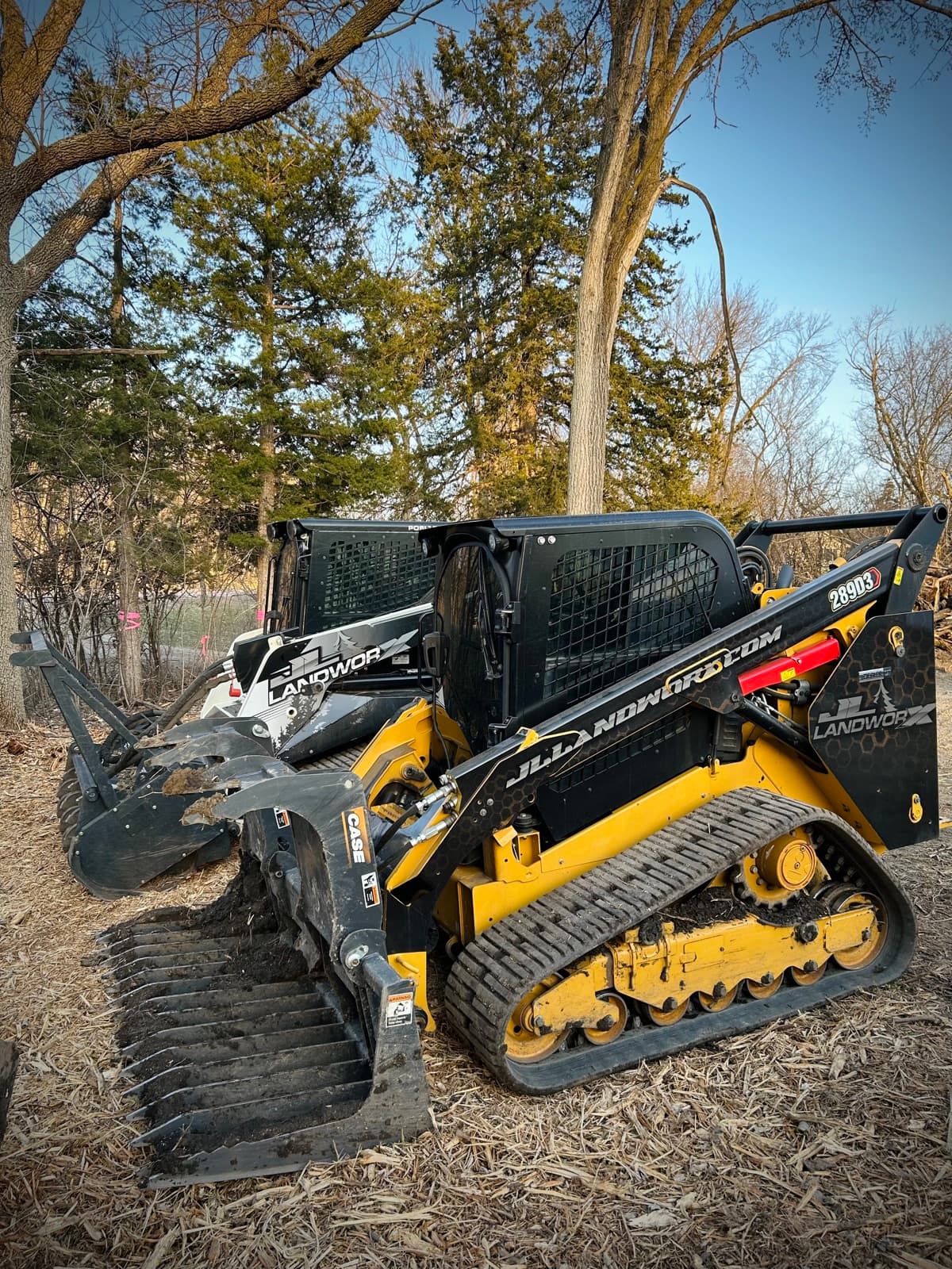 JL Landworx track loaders with rock rake and mulcher head staged in the woods between jobs