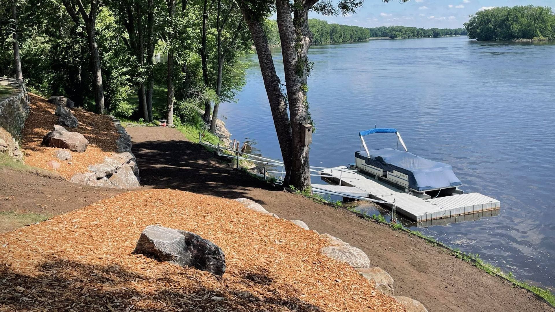 Restored riverbank with fresh mulch beds, boulder edging, and a pontoon dock on the water